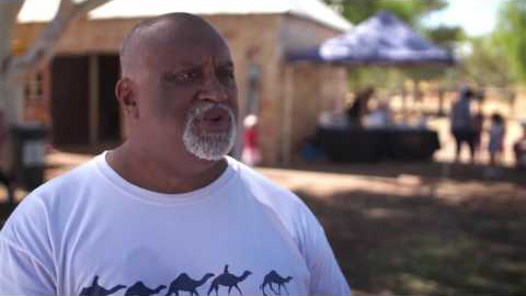An indigenous man who appears to be talking wears a white shirt with a silhouette of five camels walking on the front