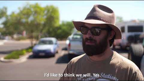 A man with a beard, wide brimmed hat and sunglasses on stands in front of the camera in a carpark as he's being interviewed. The subtitles read 