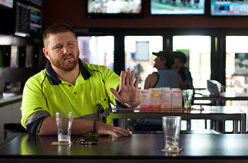 A man is sitting inside a pub wearing a high viz shirt. His sunglasses hang from his shirt pocket. On the table in front of him is two empty glasses. His keys are on the table in front of him. His left arm is raised with his palm showing in a stop signal.