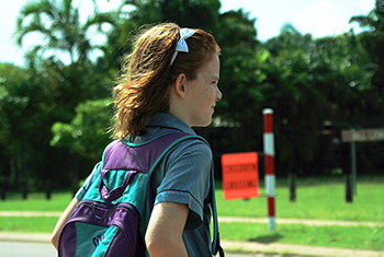 The blurred background shows a striped pedestrian crossing post and flag. Close up is the image of a school girl. She has a headband with a bow in her hair. She's in school uniform and is carrying her school backpack. The girl is standing at the edge of the path, ready to cross safely.