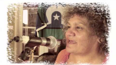 An indigenous woman sings into a microphone. She has soft curly brown hair. A Torres Strait flag hangs from the wall behind her.