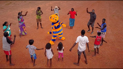 Thirteen Aboriginal peoples are standing in a circle dancing. Hector the road safety cat is standing in the middle of the circle and is also dancing.