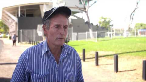 A man stands in front of a stadium as he's being interviewed. He's wearing a blue and white striped collared shirt and a navy blue hat. His mouth is slightly open as he is about to answer a question.
