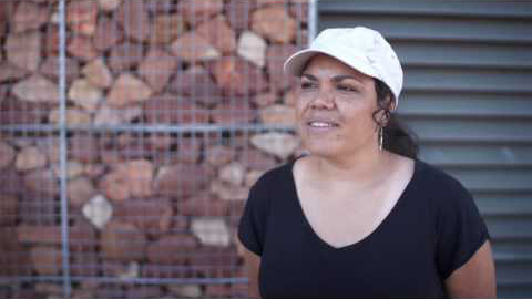 An indigenous woman stands in front of a building, She's wearing a black t-shirt and white hat. Her hands are behind her back as she is being interviewed.