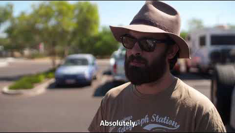 A man with a beard, wide brimmed hat and sunglasses on stands in front of the camera in a carpark as he's being interviewed. The subtitles read 
