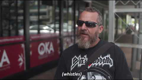 A man with a beard wearing sunglasses stands in front of the camera as he's being interviewed on a sidewalk. The subtitles read 